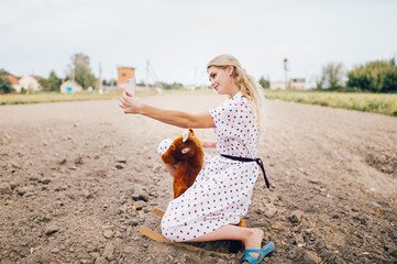 Nice looking beautiful blonde girl making selfie photo on smartphone. Young happy pretty female in stylish retro dress riding toy horse outdoor. Strange bizarre fashionable woman portrait. Childhood.