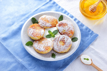 Cottage cheese pancakes sprinkled powdered sugar and fresh mint. Cheesecakes on white plate. Sour cream in wooden spoon. Honey and dairy products on white blue background. Healthy food