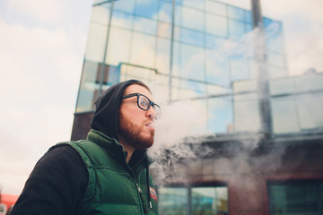 Portrait of young guy with large beard in glasses vaping an electronic cigarette opposite urban...