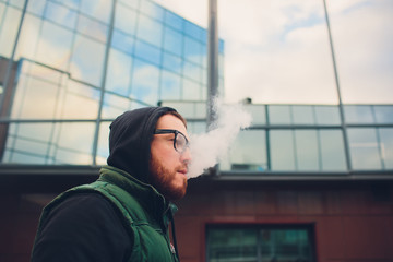 Portrait of young guy with large beard in glasses vaping an electronic cigarette opposite urban...