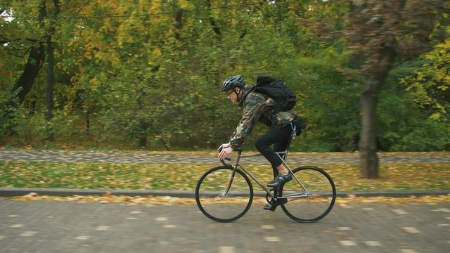 Young hipster man in helmet riding fixed gear bicycle in the city during autumn morning, side view