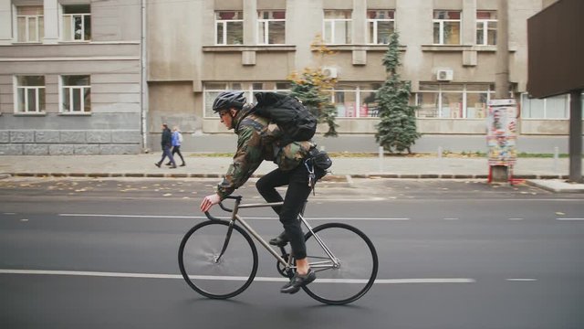 Young hipster man in helmet riding fixed gear bicycle in the city during autumn morning, side view