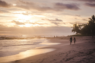 Couple walks holding hands along the long sandy paradise beach of Playa del Carmen, Santa Teresa Costa Rica during a colorful sunset