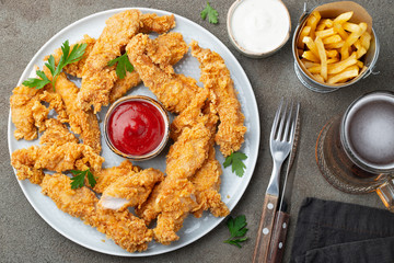 Breaded chicken strips with two kinds of sauces, fried potatoes and beer on a wooden Board. Fast food on dark brown background. Top view