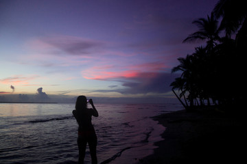Fototapeta premium Silhouette of solo female backpacker takes a photo of a sunset using her phone on a paradise beach in Central America