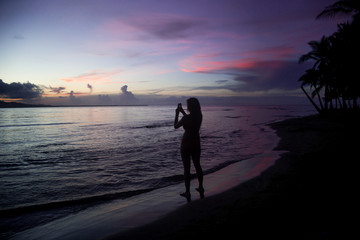 Silhouette of solo female backpacker takes a photo of a sunset using her phone on a paradise beach in Central America