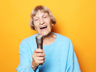 Happy old senior woman singing with microphone, having fun, expressing musical talent over yellow...