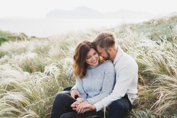 Happy young loving couple sitting in feather grass meadow, laughing and hugging, casual style sweater and jeans - 231612595