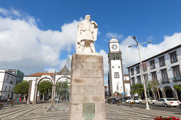 Fototapeta na wymiar Main square of Ponta Delgada with statue of Gonzalo Velho Cabral in Azores, Portugal. Saint Sabastian church with a clock tower and the historic town gates under the blue sly with cumulus clouds.