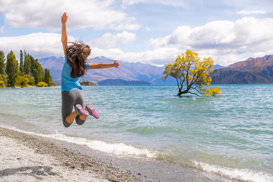 New Zealand travel happy tourist woman jumping of joy at Wanaka lake landscape with lone tree, famous attraction.