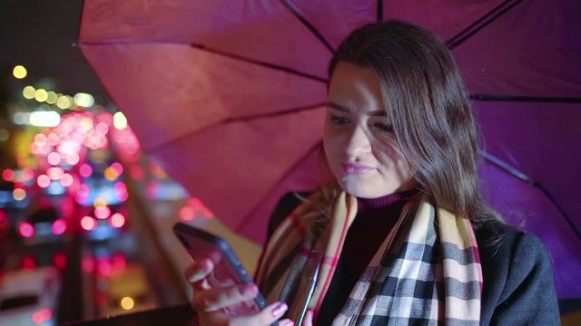 pretty young girl stands at night in the city against the background of the lights of passing cars with an umbrella in her hand hiding from the rain and writes a message on the mobile phone screen