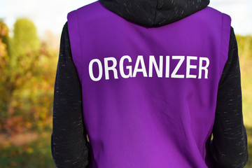 Organizer written on violet vest back of young man participate as volunteer in outdoor sport event. Blurred autumn landscape background.