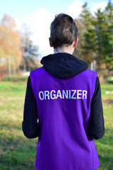 Organizer written on violet vest back of young man participate as volunteer in outdoor sport event. Blurred autumn landscape background.