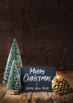 Merry Christmas and happy new year on blackboard with xmas tree and gold pine cone and snow falling on grunge wood table and dark brown wooden wall.winter holiday greeting card.