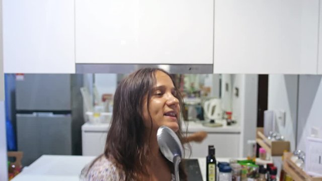 Funny pretty brunette woman housewife in pajamas cookes dances and sings song with ladle while cooking in modern kitchen at home. 4k, 3840x2160