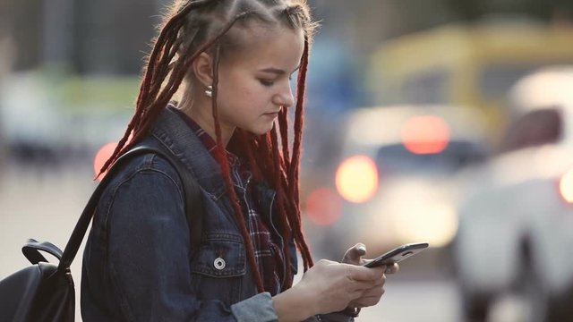 Cute girl with red dreadlocks using phone outdoors, having short break as coming back home after work