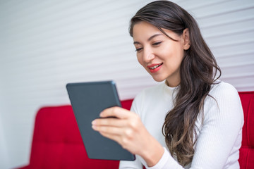 Beautiful attractive young Asian woman using smart phone or cellphone or tablet while sitting on red sofa at living room in the morning  