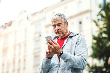 A portrait of an active mature man standing outdoors in city, using smartphone.