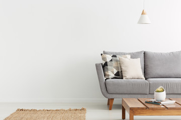 Wooden table in front of grey sofa with cushions in white flat interior with copy space. Real photo