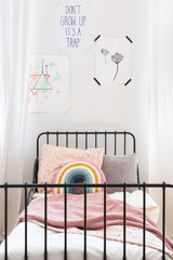 Rainbow cushion and pink blanket on child's bed in white bedroom interior with drawings. Real photo