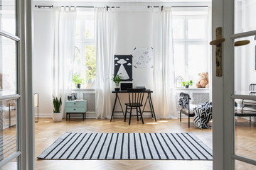 Striped carpet near black chair at desk in child's room interior with windows, bed and plants. Real...
