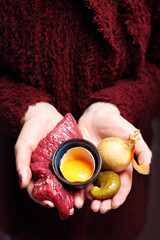 Surowe czerwone mięso. Kobieta trzyma w dłoni wołowinę, ogórki , cebulę i żółtko