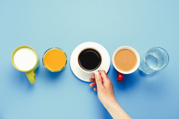 Female hand holding a cup with black coffee and group of healthy drinks, orange juice, cappuccino, water, yogurt on a blue background. Flat Lay Still Life Table Top View