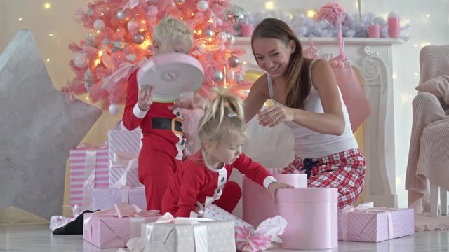 Young mother and her kids with Christmas gifts. They sit in the living room, next to the fireplace, near the elegant Xmas tree. Children in Santa Claus costumes open boxes of presents. Everyone has
