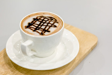 The white cup of hot capuchino and chocolate syrup on top on the wooden table background.