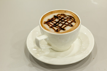 The white cup of hot capuchino and chocolate syrup on top on the wooden table background.