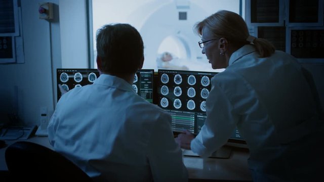 In Control Room Doctor and Radiologist Discuss Diagnosis while Watching Procedure and Monitors Showing Brain Scans Results, In the Background Patient Undergoes MRI or CT Scan Procedure.