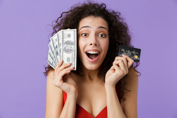 Image of delighted woman 20s wearing red dress holding fan of money and credit card, standing...