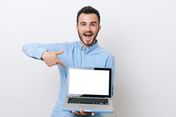 Amazed young man in casual pointing at blank screen on laptop over white background, copyspace