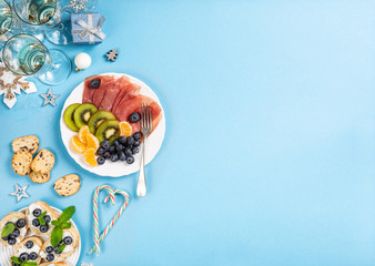 Christmas composition with meat, fruit, champagne and decorations on blue. Holidays food background with copy space for text. Top view. Flat lay.