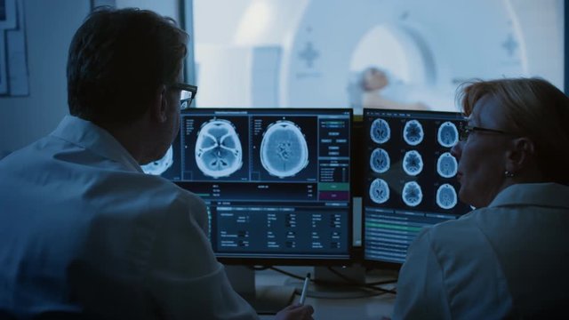 In Control Room Doctor and Radiologist Discuss Diagnosis while Watching Procedure and Monitors Showing Brain Scans Results, In the Background Patient Undergoes MRI or CT Scan Procedure.
