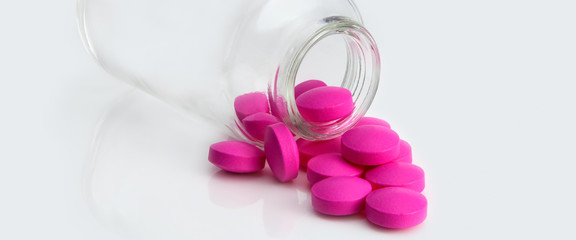 Bright pink tablets scattered from a glass jar on a gray background. Side view.