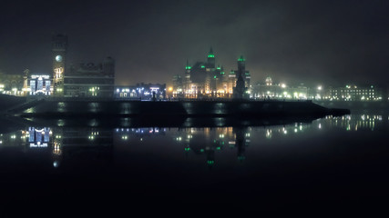 Fototapeta na wymiar mystical towers and castles of the night city illuminated by lights in ominous fog are reflected symmetrically in the cold river