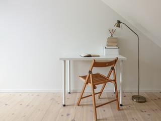 Simple workspace interior. Open book in minimalist home office room. Lamp, books, wooden floor and folding chair. Empty white wall and desk for copy space.