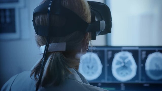 Futuristic Concept: In Medical Control Room Female Doctor Wearing Virtual Reality Headset Monitors Patient Undergoing MRI or CT Scan Procedure. Computer Displays Shows Brain Scans.