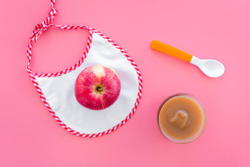 Baby food concept. Apple puree in bowl near bib, apple, spoon on pink background top view