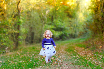 Cute little toddler girl making a walk through autumn park. Happy healthy baby enjoying walking with parents. Sunny warm fall day with child. Active leisure and activity with kids in nature.