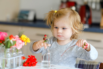 Cute beautiful little toddler girl making flower bouquet. Adorable baby child playing with different flowers. Healthy daughter, childhood and development concept. Happy kid at home or nursery.