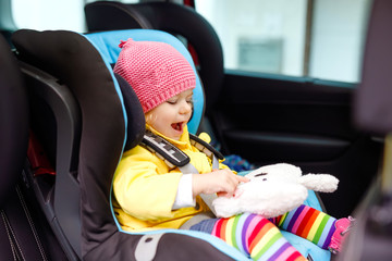 Adorable baby girl with blue eyes and in colorful clothes sitting in car seat. Toddler child in...
