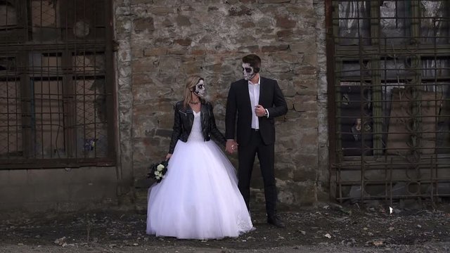A horrible couple in a wedding dress and costume with makeup in the form of a skeleton stand against an old brick wall in an old abandoned house. The concept of Halloween. Halloween face art.