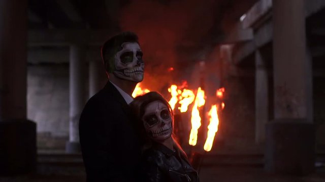 Couple in wedding clothes and with a terrible makeup for Halloween stand against a burning fire in the dark among the smoke. Halloween loving couple with skull make-up. Halloween or horror theme.