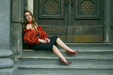 Attractive slim woman in a red elegant blouse and black skirt posing sitting on the stairs. Fashion photo