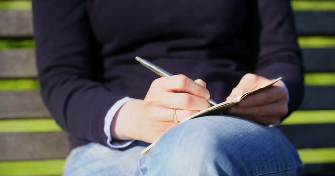 Medium closeup shot of a young Caucasian woman writing notes on a paper notepad in a park.