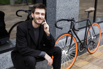 Fototapeta na wymiar Photo of stylish man 20s using cell phone, while sitting outdoor with bicycle
