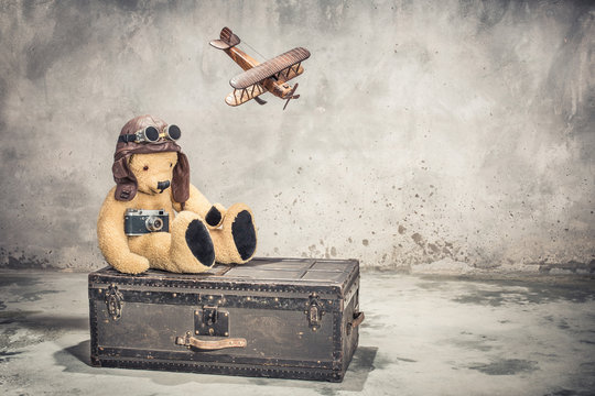 Teddy Bear with leather aviator's hat and goggles sitting on vintage old aged classic travel trunk circa 1900s with film camera and flying toy plane. Travel by air concept. Retro style filtered photo