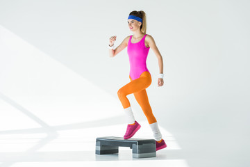 Fototapeta na wymiar smiling sporty woman exercising on step platform and looking away on grey